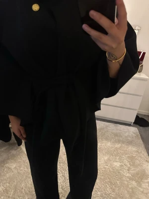 Svart kappa med knytband och guldknapp - Stilren svart poncho kappa med knytband i midjan och en dekorativ guldig knapp vid halsen. Kappan har vida ärmar och en elegant, rak passform. Perfekt för dig som gillar trendig stil, kappan är väldigt tunn och skulle passa för sena utekvällar på sommaren och våren. Den är använd 1-2 gånger max.