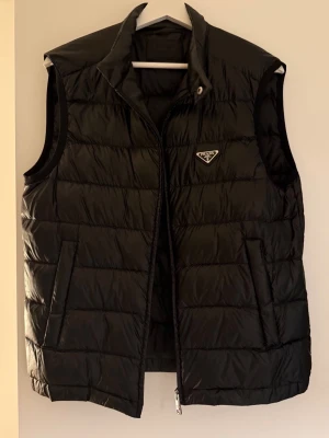 Svart dunväst från Prada - Snygg svart dunväst från Prada med quiltad design och hög krage. Västen har dragkedja framtill, två fickor och en diskret logotyp på bröstet. Perfekt för lager-på-lager och ger en stilren look. Storleken är 50 men motsvarar L