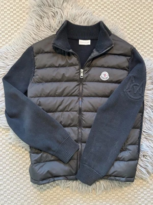 BYTES! - Snygg svart cardiganjacka från Moncler med quiltad front och stickade ärmar. Jackan har dragkedja framtill, ribbad krage och muddar samt Moncler-logga på bröstet och diskret broderad logga på ärmen. Perfekt för lager på lager och stilrena outfits.