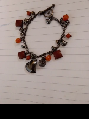 Armband med hjärthänge och orange pärlor - Charmigt armband i mörkmetall med flera hängen, bland annat ett hjärta, små kulor och orangea pärlor i olika former. Kedjan har ett klassiskt stavlås och dekoreras av både runda och fyrkantiga detaljer. Perfekt accessoar för att ge din stil en unik touch.