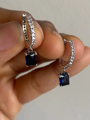 Pandora Silverörhängen med blå sten och kristaller - Snygga örhängen i silverfärg med små gnistrande kristaller på ringen och en rektangulär mörkblå sten som hänger ned. Perfekt för dig som gillar stilrena och eleganta accessoarer med lite extra bling.material: s925 sterling silver and zirconia sten