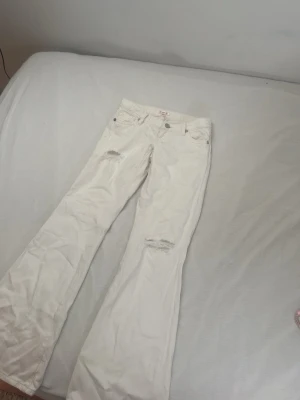 Vita bootcut/loose med slitningar - Säljer ett par vita bootcut jeans med slitna detaljer på låret och knät. Jeansen har klassisk femficksdesign, hällor för skärp och stängs med knapp och dragkedja. Perfekta för en trendig och avslappnad look.