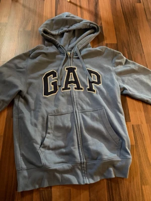 Blå GAP hoodie med dragkedja - Snygg blå hoodie från GAP med dragkedja framtill och stor logga över bröstet. Tröjan har huva med dragsko och två stora fickor framtill. Perfekt för en avslappnad och cool stil. Mjuk bomullsblandning som är skön att ha på sig.