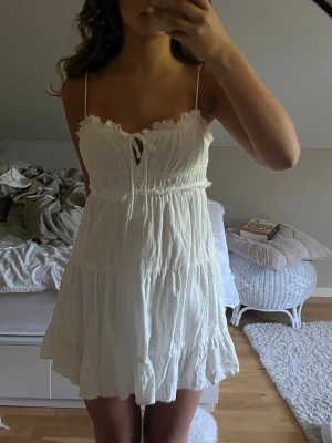 Vit kort sommarklänning med volang - Söt vit kort klänning med tunna axelband, volangkant upptill och nertill samt knytning framtill. Klänningen har en luftig passform och är tillverkad i ett lätt bomullsliknande material, perfekt för varma dagar.