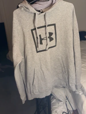 Grå hoodie från Under Armour - Snygg grå hoodie från Under Armour med svart logga på bröstet. Klassisk modell med huva och känguruficka framtill. Mjuk bomullsblandning och ribbade muddar vid ärmslut och nederkant. Perfekt för en avslappnad streetstyle.