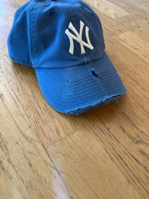 Blå New York Yankees keps - Snygg blå keps från 47 Brand med New York Yankees-logga broderad framtill i vitt. Kepsen har böjd skärm med slitna detaljer och hål, samt justerbar passform. Tillverkad i bomull.