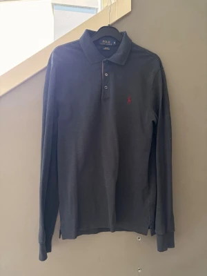 Mörkblå långärmad piké från Polo Ralph Lauren - Snygg marinblå långärmad pikétröja från Polo Ralph Lauren med klassisk krage och knappar framtill. Tröjan har en liten röd broderad logga på bröstet och är tillverkad i mjuk bomull. Bara att höra av sig vid eventuella frågor!😉