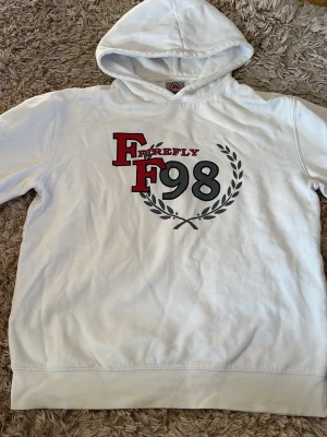 Vit hoodie från Firefly med tryck - Snygg vit hoodie från Firefly med stor huva och tryck framtill med texten 'FF98' och 'Firefly' i rött och svart. Dekorativ lagerkrans runt siffrorna ger en sportig vibe. Perfekt för dig som gillar streetwear och vill sticka ut med en unik design.