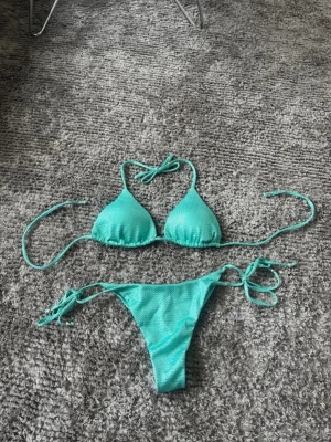 Turkos bikini med knyt och shimmerig - Snygg turkos bikini med trekantsöverdel och knyt i både rygg och nacke. Underdelen har knyt i sidorna och båda delarna har en lätt meshstruktur som ger en cool look. Perfekt för stranden eller poolen i sommar! Aldrig använd!!! Nypris 800kr