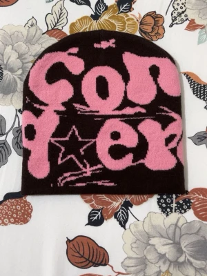 Brun och rosa mössa med grafiskt tryck - Snygg mössa i brunt med stort rosa grafiskt tryck och stjärndetalj. Mjuk och stretchig stickning, perfekt för att addera lite edge till din look. Passar dig som gillar streetwear och statement-plagg.