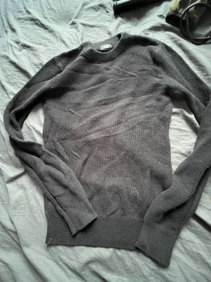 Mörkgrå stickad tröja - En enkel och stilren mörkgrå stickad tröja med rund halsringning och ribbade muddar vid ärmslut och nederkant. Tröjan har en klassisk passform och är perfekt för lager på lager. Materialet känns mjukt och bekvämt mot huden. Den har ett litet hål som knappt märks när man har tröjan på sig och som är lätt att fixa om man väl kan det.