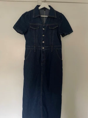 Mörkblå jeansklänning med knappar - Snygg lång jeansklänning i mörkblå denim med korta ärmar, krage och knappar framtill. Klänningen har två bröstfickor och markerad midja för en cool vintagekänsla. Perfekt för dig som gillar klassisk denimstil. Stl L men är mer en S