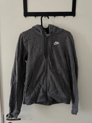 Hoodie från Nike - Mörkgrå hoodie från Nike med dragkedja framtill och vit broderad logga på bröstet. Passar stl. Xs- S