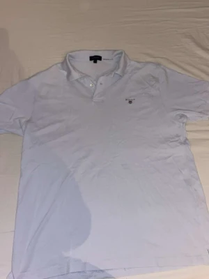 Vit piké från Gant - Klassisk vit pikétröja från Gant med liten logga broderad på bröstet. Tröjan har korta ärmar, krage och knappar vid halsen. Perfekt till en avslappnad och stilren look.