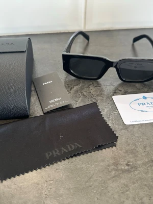 Svarta solglasögon från Prada - Snygga svarta solglasögon från Prada med rektangulära bågar och diskret logga på sidan. Kommer med originalfodral, putsduk och äkthetskort. Perfekt accessoar för en trendig look.