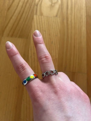 Två coola ringar i silverfärg - Säljer två ringar: en i silverfärg med utskurna stjärnor runt om och en annan i silverfärg med färgglada prickar i blått, grönt, gult och rött. Båda ringarna har en unik design och passar perfekt för dig som vill sticka ut.