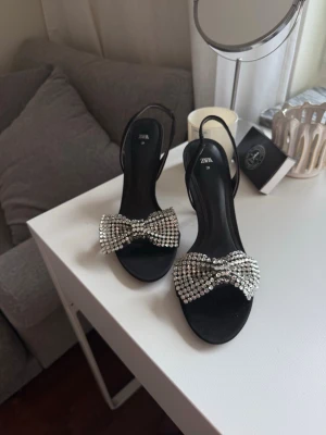 Svarta pumps med strassrosett  - Säljer ett par svarta pumps från Zara med öppen tå och smal klack. Skorna har en stor glittrig rosett framtill täckt av strass. 10 cm höga. Finns ej längre i butik.