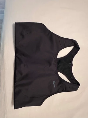 Svart sport-bh från Nike Dri-FIT - Snygg svart sport-bh från Nike med Dri-FIT-material som andas. Klassisk racerback-design och diskret Nike-logga framtill. Perfekt för träning och ger bra stöd. Materialet är stretchigt och bekvämt för aktiva dagar.