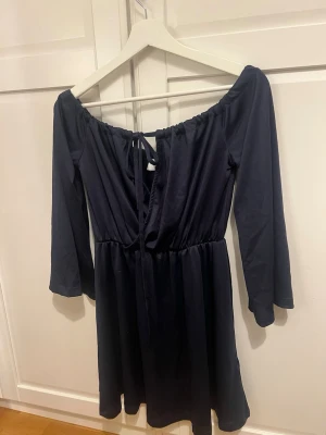 Marinblå offshoulder klänning Linn Ahlborg - Snygg marinblå offshoulder klänning från Linn Ahlborg x Rebecca Stella. Klänningen har långa ärmar, resår i midjan och en söt knytning framtill. Perfekt för dig som gillar en enkel men trendig look.
