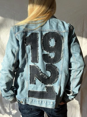 Jeansjacka med stora siffror - Snygg ljusblå jeansjacka med stora svarta siffror '1921' i patchad design på ryggen. Perfekt statement-plagg för dig som vill sticka ut🩵