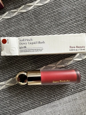 Soft Pinch Dewy Liquid Blush Worth - Säljer en Soft Pinch Dewy Liquid Blush från Rare Beauty i färgen Worth. Rougen är flytande och kommer i en lyxig matt rosa tub med guldfärgat lock. Perfekt för att ge kinderna en naturlig och fräsch look. Innehåller 7,5 ml.