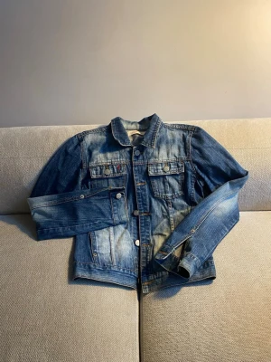 Klassisk blå jeansjacka - Snygg blå jeansjacka med slitna detaljer och två bröstfickor med knappar. Jackan har silverfärgade knappar framtill och vid ärmsluten samt justerbara spännen i midjan. Perfekt oversized look och tidlös design som passar till allt.