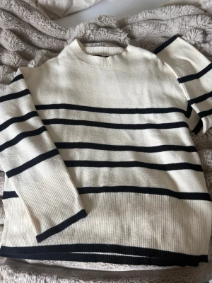 Randig stickad tröja i vitt och svart - Säljer en snygg stickad tröja i vitt med svarta horisontella ränder. Tröjan har rund halsringning, långa ärmar och ribbade muddar samt nederkant. Materialet känns mjukt och bekvämt, perfekt för en avslappnad look.