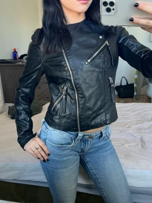 Svart biker skinnjacka med dragkedjor - Snygg svart skinnjacka i bikerstil med flera silvriga dragkedjor framtill och figurnära passform. Jackan har en cool asymmetrisk design och ger en edgy look till din outfit. Perfekt för dig som vill ha en klassisk men ändå trendig skinnjacka.