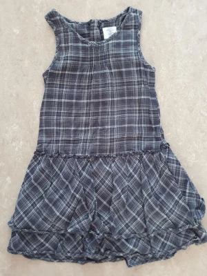 Klänning - Säljer en grå ärmlös klänning från H&M med rutigt mönster och volangkant nedtill. Klänningen har en avslappnad passform och är tillverkad i mjuk bomull, perfekt för varma dagar.