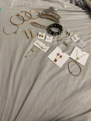 Mix av smycken i guld - Säljer ett blandat paket med smycken: flera armband och halsband i guld- och silverfärg, örhängen i olika former, samt ringar. Några smycken har pärlor och stenar, och det finns även ett svart armband med nitar. Perfekt för dig som vill variera din stil.