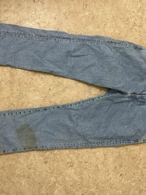 Ljusblå raka jeans - Säljer ett par ljusblå jeans med rak passform och klassisk femficksdesign. Jeansen har en patchad detalj på ena benet och är tillverkade i denim. Perfekta för en avslappnad och trendig look.