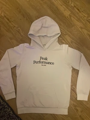 Vit hoodie från Peak Performance - Snygg vit hoodie från Peak Performance med svart logga på bröstet och klassisk känguruficka. Mjuk insida och ribbade muddar vid ärmar och midja. Perfekt för en avslappnad och stilren look.