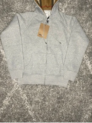 Grå zip hoodie från Burberry - Snygg grå zip hoodie från Burberry med klassiskt rutigt foder i huvan och broderad logga på bröstet. Hoodien har dragsko i huvan, två fickor framtill och ribbade muddar. Perfekt för en avslappnad men stilren look.