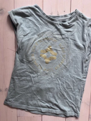 Ljusblå t-shirt från Sofie Schnoor - T-shirt i ljusblått från Sofie Schnoor med ett stort guldfärgat tryck på bröstet. Modellen är ärmlös med rund halsringning och har en avslappnad passform. Perfekt för dig som gillar en enkel men ändå snygg look.