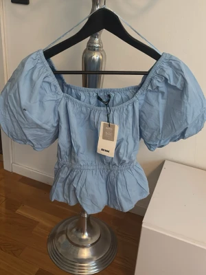 Ljusblå puffärm topp från Bik Bok - Supersöt ljusblå topp från Bik Bok med puffiga korta ärmar och resår i midjan för en feminin siluett. Toppen har en luftig och croppad passform, perfekt till sommaren. Tillverkad i mjuk bomull och har en söt volangkant nedtill.