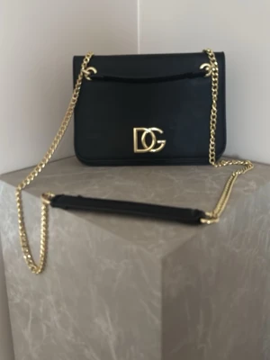 Svart axelväska från Dolce & Gabbana - Snygg svart axelväska i slätt läder från Dolce & Gabbana med guldfärgad kedjerem och DG-logga framtill. Väskan har en stilren, rektangulär form och minimalistisk design som ger en lyxig känsla. Perfekt för dig som gillar exklusiva accessoarer. Bredd: 23 cm och 16 cm hög 