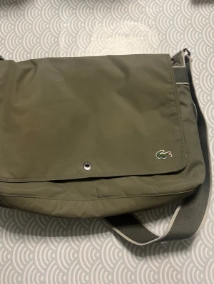 Olivgrön axelväska från Lacoste - Snygg olivgrön axelväska från Lacoste med klassisk krokodillogga på framsidan. Väskan har justerbar axelrem i matchande färg och är tillverkad i slitstarkt syntetmaterial. Perfekt för dig som vill ha en stilren och praktisk väska till vardagen.