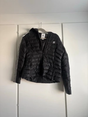 Svart dunjacka med huva från Adidas - Svart dunjacka från Adidas med huva och dragkedja framtill. Klassiska vita ränder längs ärmarna och Adidas-logga på bröstet. Jackan har en sportig look och är lätt vadderad för extra värme. Perfekt för kalla vinterdagar. Pris kan diskuteras💕Skulle även säga att den är liten i storleken