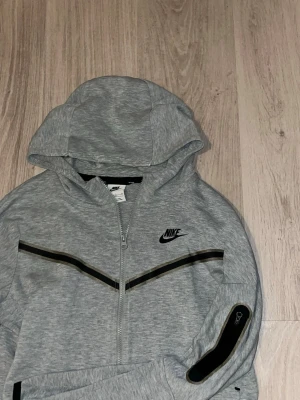Grå Nike tech flece - Snygg grå hoodie från Nike med dragkedja framtill och svart Nike-logga på bröstet. Hoodien har svarta detaljer längs bröstet och ärmen samt en praktisk huva. Perfekt för en sportig och avslappnad stil.