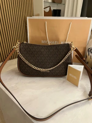 Brun axelremsväska från Michael Kors - Snygg brun handväska från Michael Kors med klassiskt MK-mönster, guldfärgad kedjedetalj och justerbar axelrem i läder. Väskan har en elegant, avlång form och dragkedja upptill. Perfekt accessoar för att lyfta din outfit.