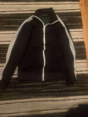Svart vindjacka med vita detaljer - Sportig svart vindjacka med vita ränder längs ärmarna och vit dragkedja framtill. Jackan har hög krage och lång ärm, perfekt för en avslappnad streetwear-look.