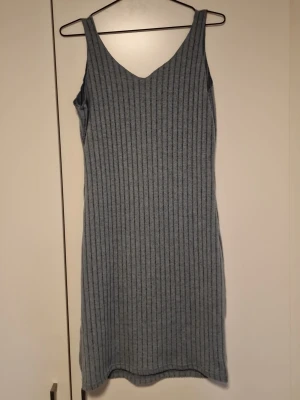 Grå ribbad ärmlös klänning - Säljer en enkel och stilren grå ribbad klänning med v-ringning och ärmlös design. Klänningen är i mjukt stickat material och har en kroppsnära passform som framhäver siluetten. Perfekt för dig som gillar minimalistisk stil.
