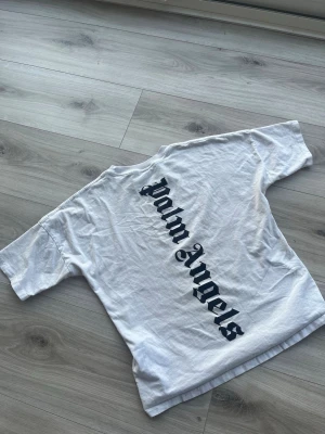 Äkta Palm Angels T-shirt - Säljer en vit t-shirt från Palm Angels med stort svart tryck i gotisk stil på ryggen och mindre tryck framtill. Klassisk rund hals och korta ärmar. Perfekt för dig som gillar streetwear och vill sticka ut med en ikonisk logga.