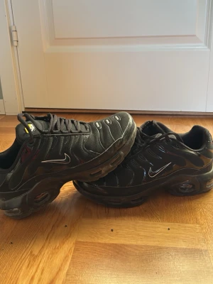 Nike Air Max Plus svarta sneakers - Nike Air Max Plus sneakers i helsvart med vita detaljer och ikonisk Swoosh på sidan. Ovandelen är i mesh och syntet med vågiga linjer och synlig Air-dämpning i sulan. Skorna har snörning och en sportig, chunky siluett som sticker ut.