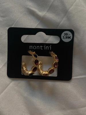 Guldiga örhängen med färgade stenar - Säljer ett par chunky guldiga örhängen från Montini med flera färgade stenar i rött, blått och rosa. Örhängena har en halvcirkelformad design och detaljerad struktur som ger en lyxig känsla. Perfekta för att addera färg till din look.