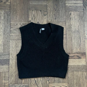 Svart stickad väst från H&M Divided - Säljer en svart stickad väst från H&M Divided. Västen har djup v-ringning, bred ribbstickad kant och är ärmlös. 