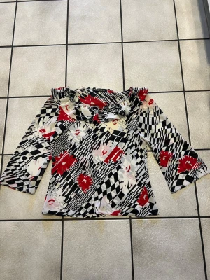 Mönstrad offshoulder blus från Berreta storlek M/L - Säljer en unik offshoulder blus från Berreta med volang vid halsen. Blusen har ett grafiskt mönster i svart, vitt och rött samt blommiga detaljer. Materialet känns lätt och flowy, perfekt för att sticka ut i mängden.