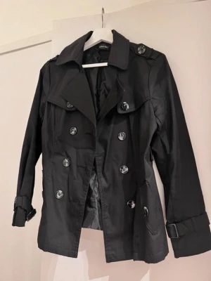 Svart trenchcoat från Vero Moda - Stilren svart trenchcoat från Vero Moda med dubbelknäppning, bred krage och bältesdetaljer vid ärmsluten. Jackan har stora knappar och två fickor framtill. Materialet är glansigt och ger en modern look.