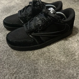 2022 Travis phantom low - Svarta Nike Air sneakers i low-modell med kontrastsömmar i vitt. Ovandelen är i mocka och textil, med klassisk swoosh på sidan och perforerad tåbox. Skorna har svart yttersula och svarta skosnören för en clean look.