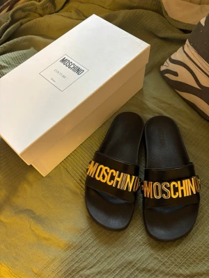 Svarta slides från Moschino - Säljer ett par svarta slides från Moschino med bred rem och stora guldfärgade bokstäver som stavar ut märkesnamnet. Tillverkade i syntetmaterial med en klassisk slip-in design. Perfekta för dig som vill ha en statement sandal till sommaren.